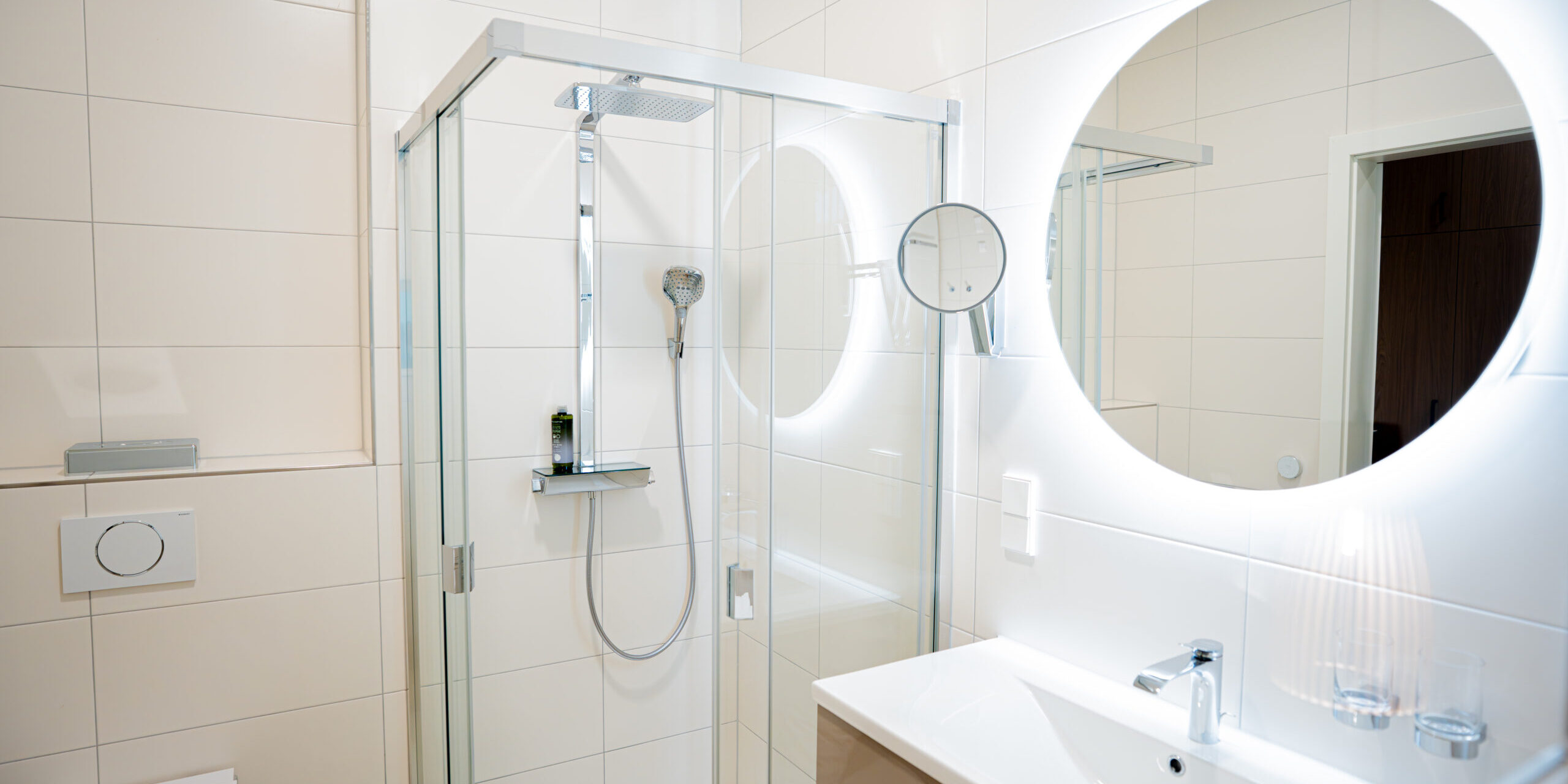 Modernes, weißes Bad mit Glasdusche, rundem Spiegel, Waschbecken und Toilette - perfekt für Ihren Aufenthalt im Komfort Doppelzimmer.