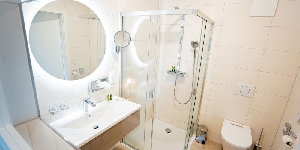 Modernes Badezimmer mit rundem Spiegel, Glasdusche, Waschbecken und wandmontierter Toilette in einem hellen, sauberen Komfort-Doppelzimmer.