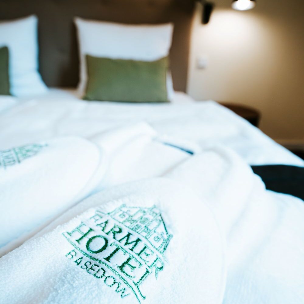 Weiße Bademäntel mit dem Logo des Farmer Hotels Basedow liegen auf einem sauber gemachten Doppelbett, das den Komfort und die einladende Note der grünen Kissen im Hintergrund unterstreicht.
