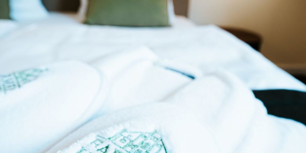 Weiße Bademäntel mit dem Logo des Farmer Hotels Basedow liegen auf einem sauber gemachten Doppelbett, das den Komfort und die einladende Note der grünen Kissen im Hintergrund unterstreicht.
