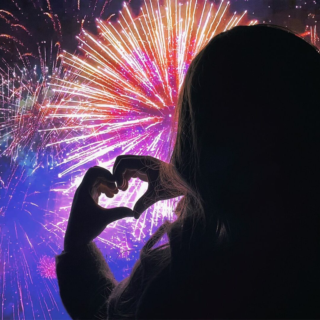Silhouette einer Person, die mit den Händen eine Herzform bildet, vor einem bunten Feuerwerk bei Nacht, das die Freude und Aufregung von Silvester 2027 einfängt.