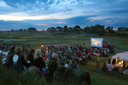 In der Abenddämmerung nehmen die Besucher des "WanderKino 2026" auf einer Wiese in Stühlen vor einer riesigen Leinwand Platz und genießen einen unvergesslichen Abend unter freiem Himmel.