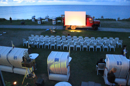 Genießen Sie mit dem Ticket "WanderKino 2026" eine magische Vorführung in der Abenddämmerung am Meer - Stuhlreihen vor einer auf einem LKW montierten Leinwand. Bringen Sie Ihr Ticket für einen unvergesslichen Filmabend unter freiem Himmel mit!.