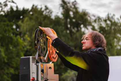 Ein Mann mit einem Ticket "WanderKino 2026" lädt Filmrollen in einen Außenprojektor, der von Bäumen umgeben ist.