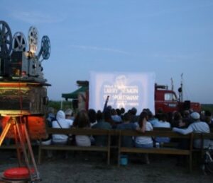 Das Publikum genießt einen Freiluftfilm in der Abenddämmerung, bei dem ein alter Projektor leuchtet - ein unvergesslicher Moment mit dem Ticket "WanderKino 2026", das die Magie des Kinos zum Leben erweckt.