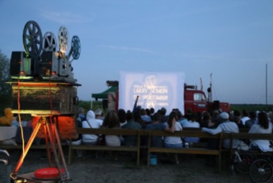Das Publikum genießt einen Freiluftfilm in der Abenddämmerung, bei dem ein alter Projektor leuchtet - ein unvergesslicher Moment mit dem Ticket "WanderKino 2026", das die Magie des Kinos zum Leben erweckt.