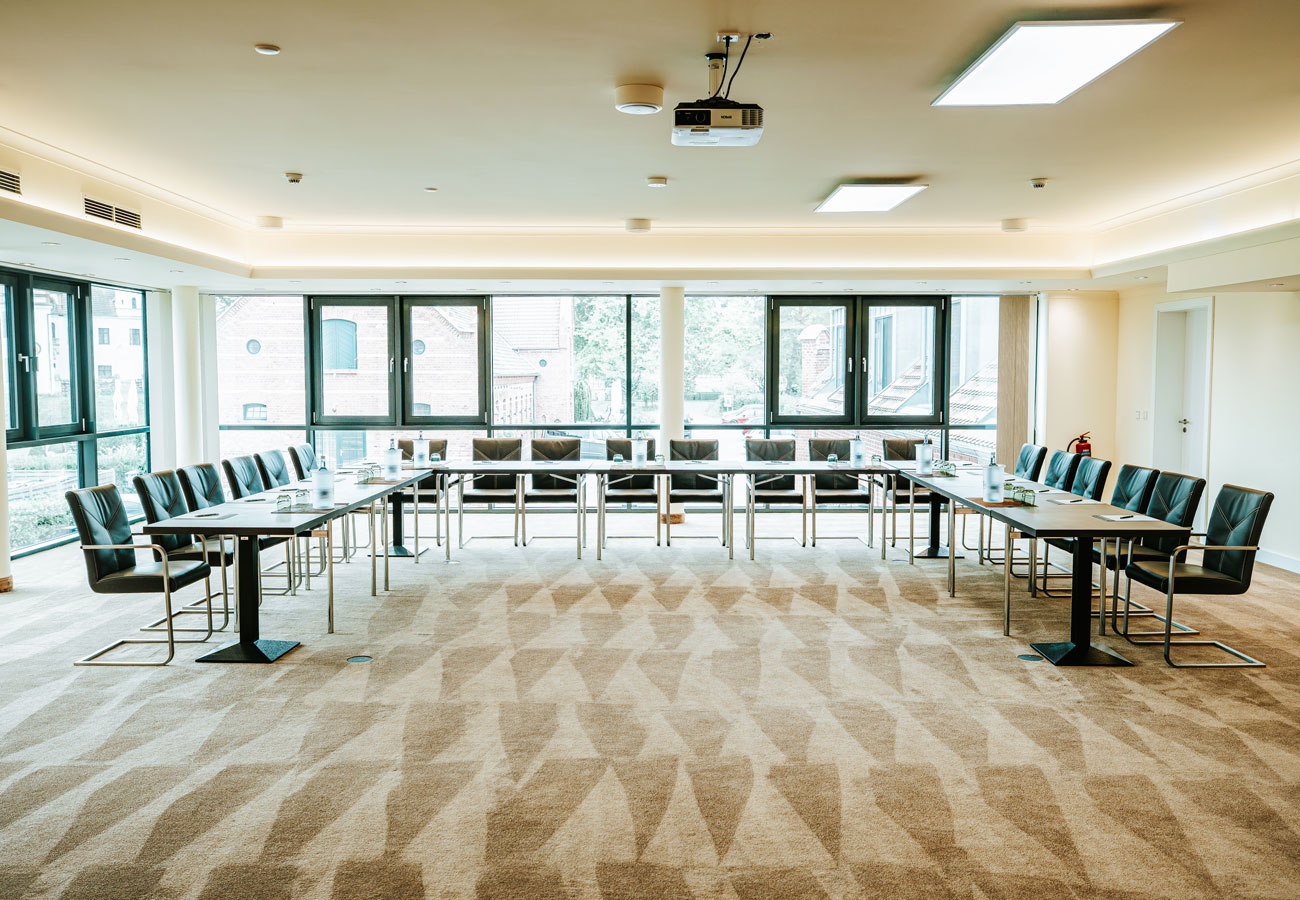 Moderner Konferenzraum mit U-förmigem Tisch, schwarzen Stühlen und großen Fenstern, die natürliches Licht hereinlassen.