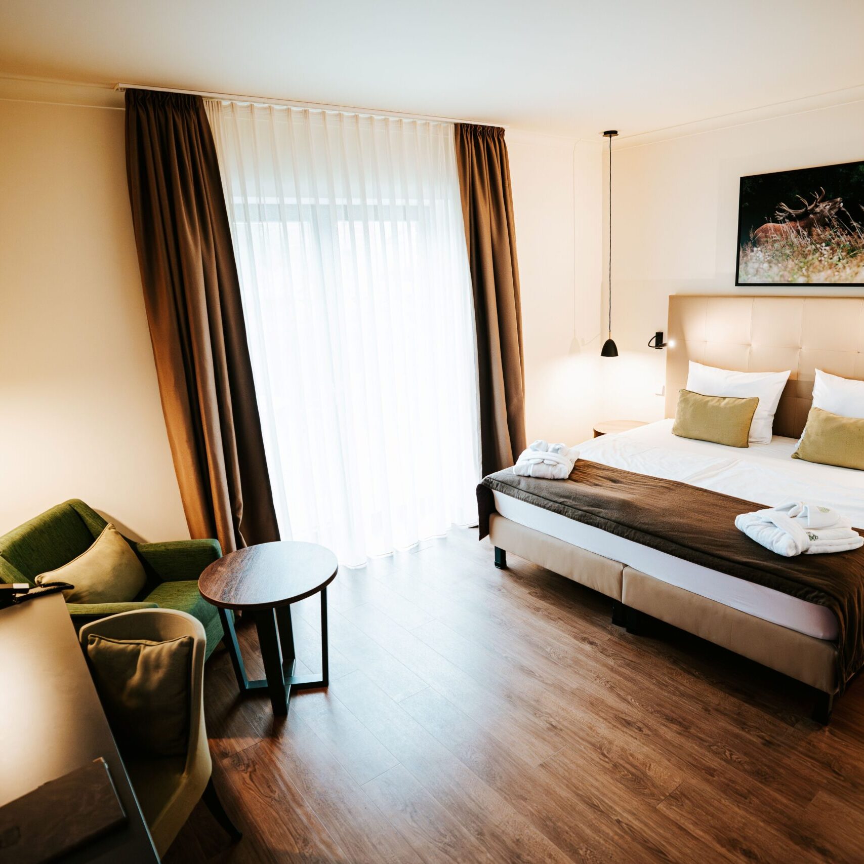 Modernes Hotelzimmer mit einem Doppelbett, einem Sessel, einem Schreibtisch und einem großen Fenster mit transparenten Vorhängen.