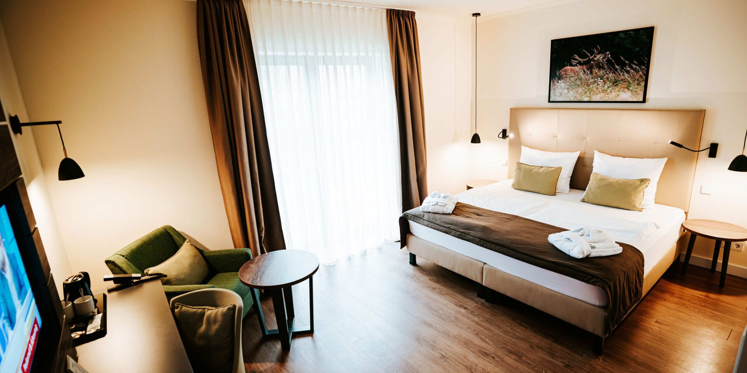 Modernes Hotelzimmer mit einem Doppelbett, einem Sessel, einem Schreibtisch und einem großen Fenster mit transparenten Vorhängen.