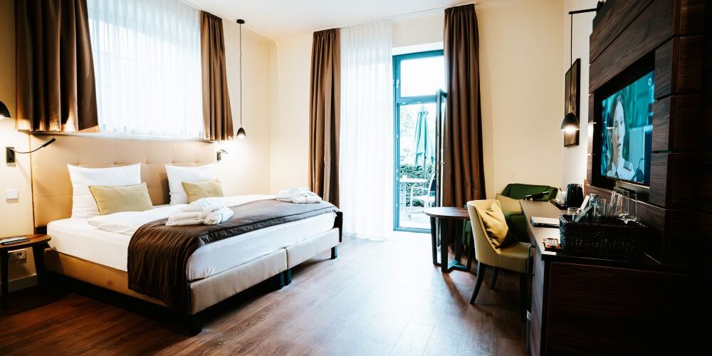 Modernes Doppelzimmer mit einem Doppelbett, Fernseher, Schreibtisch, Stühlen und großen Fenstern mit braunen Vorhängen, die einen wunderschönen Blick auf das Schloss bieten.