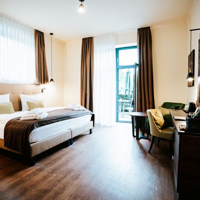 Modernes Doppelzimmer mit einem Doppelbett, Fernseher, Schreibtisch, Stühlen und großen Fenstern mit braunen Vorhängen, die einen wunderschönen Blick auf das Schloss bieten.