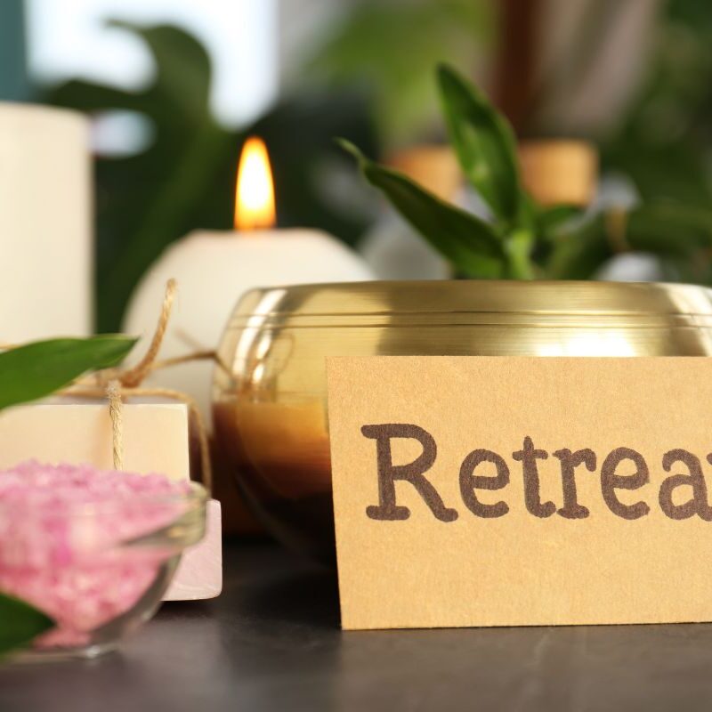 Karte mit „Retreat“ und „Glücks-Arrangement“ neben Wellness-Artikeln: Kerze, rosa Badesalz, Seife und grüne Blätter in Weichzeichneroptik.