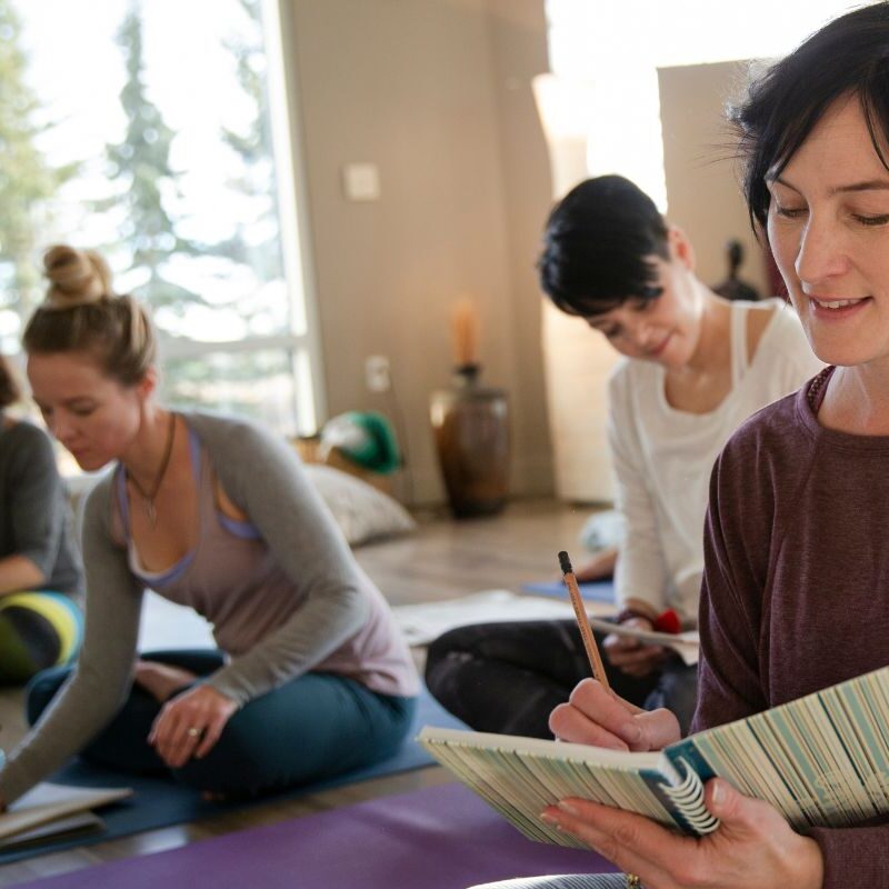Vier Frauen sitzen in einem hellen Raum auf Yogamatten und schreiben in Notizbücher, während durch das Fenster Bäume zu sehen sind – eine friedliche Szene, die die Essenz eines Glücks-Arrangements einfängt.