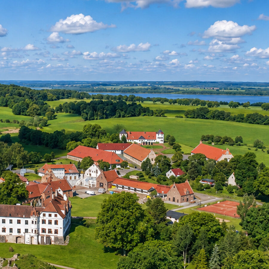 Luftaufnahme eines ländlichen Dorfes mit Gebäuden mit roten Dächern, grünen Feldern, Bäumen und einem See in der Ferne.